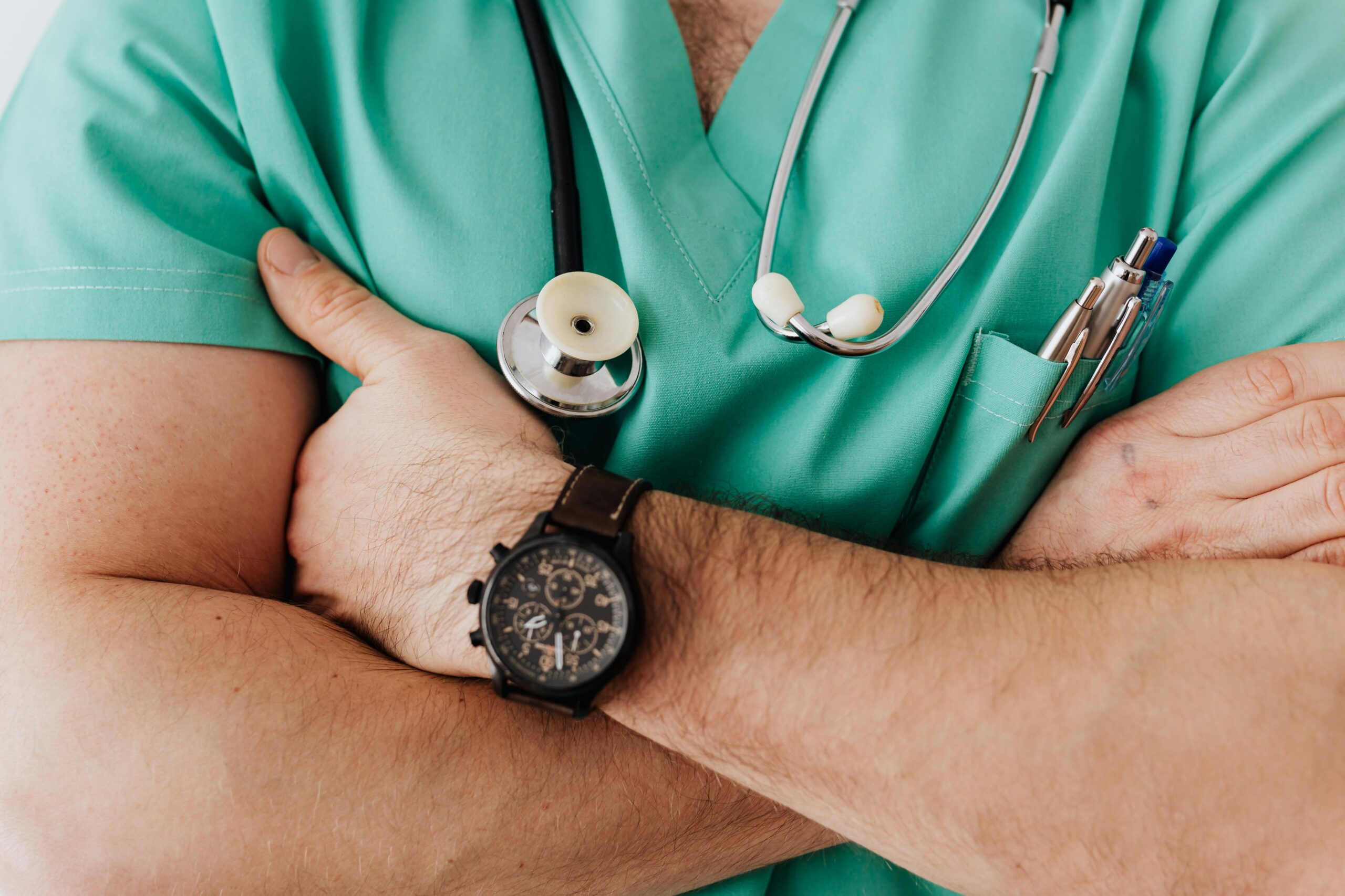 Ein medizinischer Mitarbeiter im grünen Kittel mit verschränkten Armen, einer Armbanduhr und einem Stethoskop um den Hals, der sich im MedTech-Bereich auskennt.