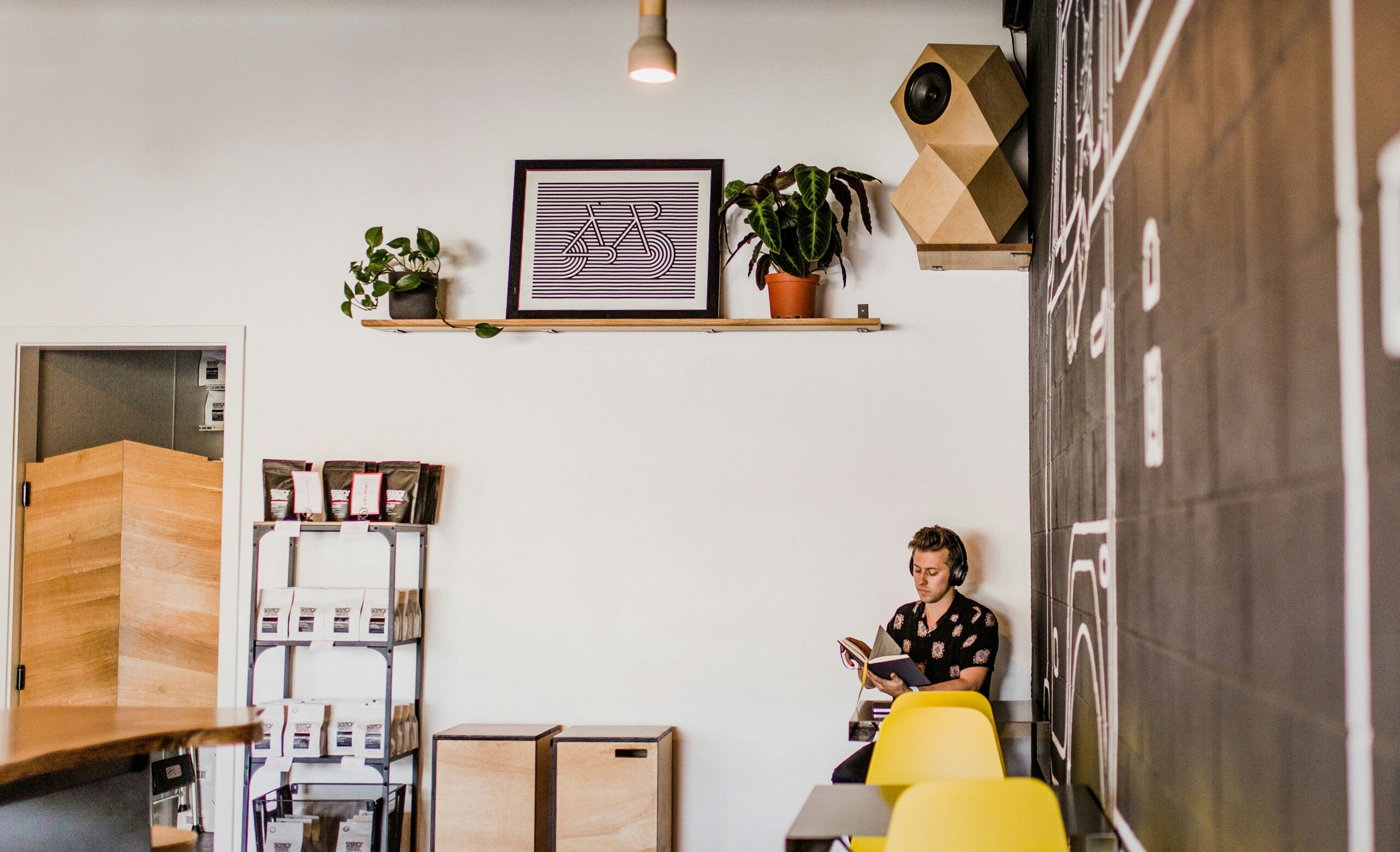 Eine Person sitzt an einem Tisch und liest ein Buch in einem modernen Café. An der Wand hängen Regale mit Pflanzen und gerahmten Kunstwerken, die an das kreative Ambiente der Bafa Unternehmensberatung erinnern. Eine Tafel mit Skizzen und leuchtend gelbe Stühle an der Theke runden das lebendige Ambiente ab.