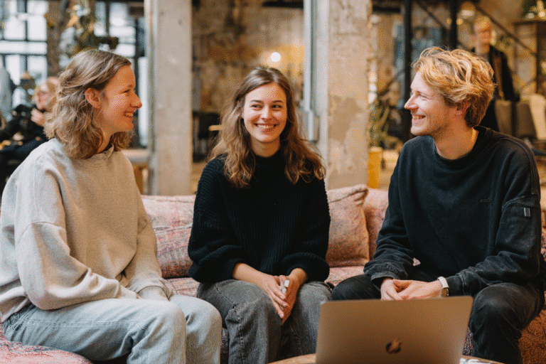 Drei junge Erwachsene sitzen auf einer Couch, lächeln und unterhalten sich, vor ihnen ein Laptop in einer gemütlichen Umgebung - perfekt für eine inspirierende Startup-Coaching-Sitzung.
