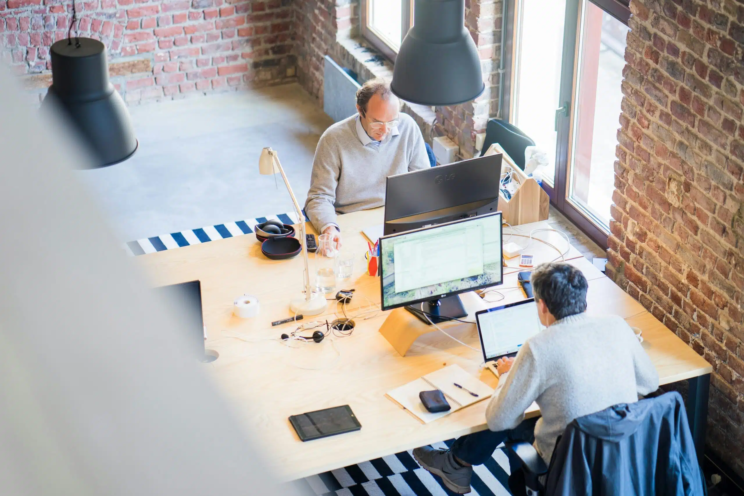 Zwei Personen, die an Schreibtischen mit Computern in einem modernen Büro mit Backsteinwänden und großen Fenstern arbeiten und mit Hilfe von CFO-as-a-Service-Lösungen gemeinsam an Finanzstrategien arbeiten.