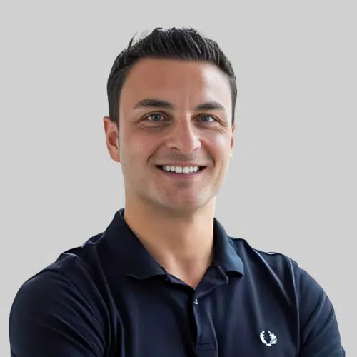 Ein Mann mit kurzen dunklen Haaren in einem marineblauen Poloshirt lächelt vor einem schlichten hellgrauen Hintergrund in die Kamera und verkörpert damit „Erfolgsbeispiele“.